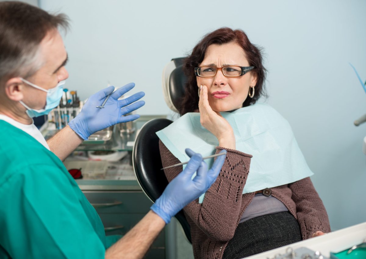 Woman feeling toothache, touching cheek with hand at dental clinic. Senior dentist trying to help. Dental care and health concept