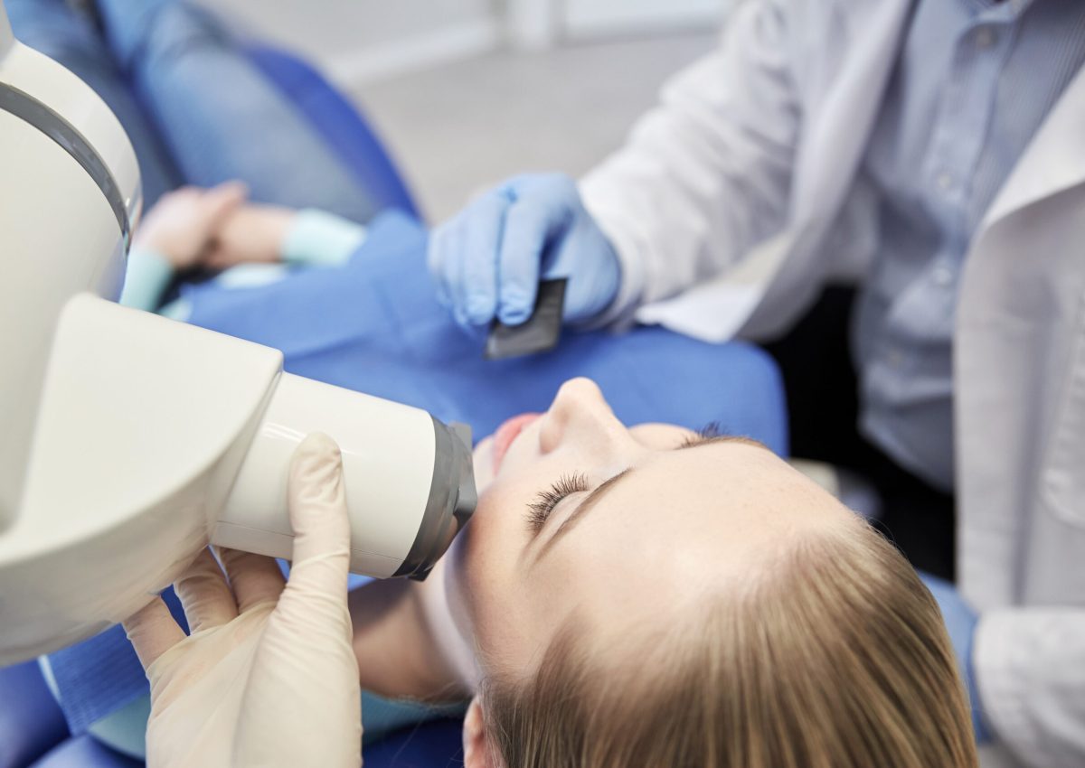 people, medicine, stomatology, technology and health care concept - close up of dentist putting intraoral shield to female patient mouth and assistant directing x-ray machine at dental clinic